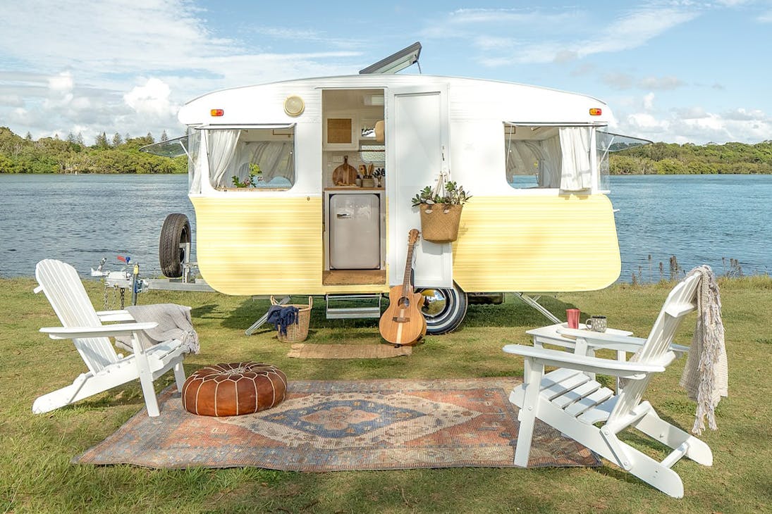 a vintage yellow caravan with white chairs out front by a river